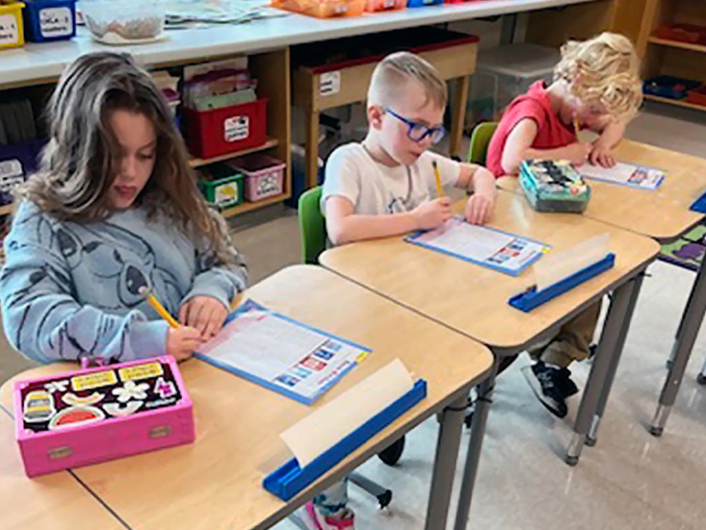 Three young students sit at desks in a classroom and write on worksheets with pencils