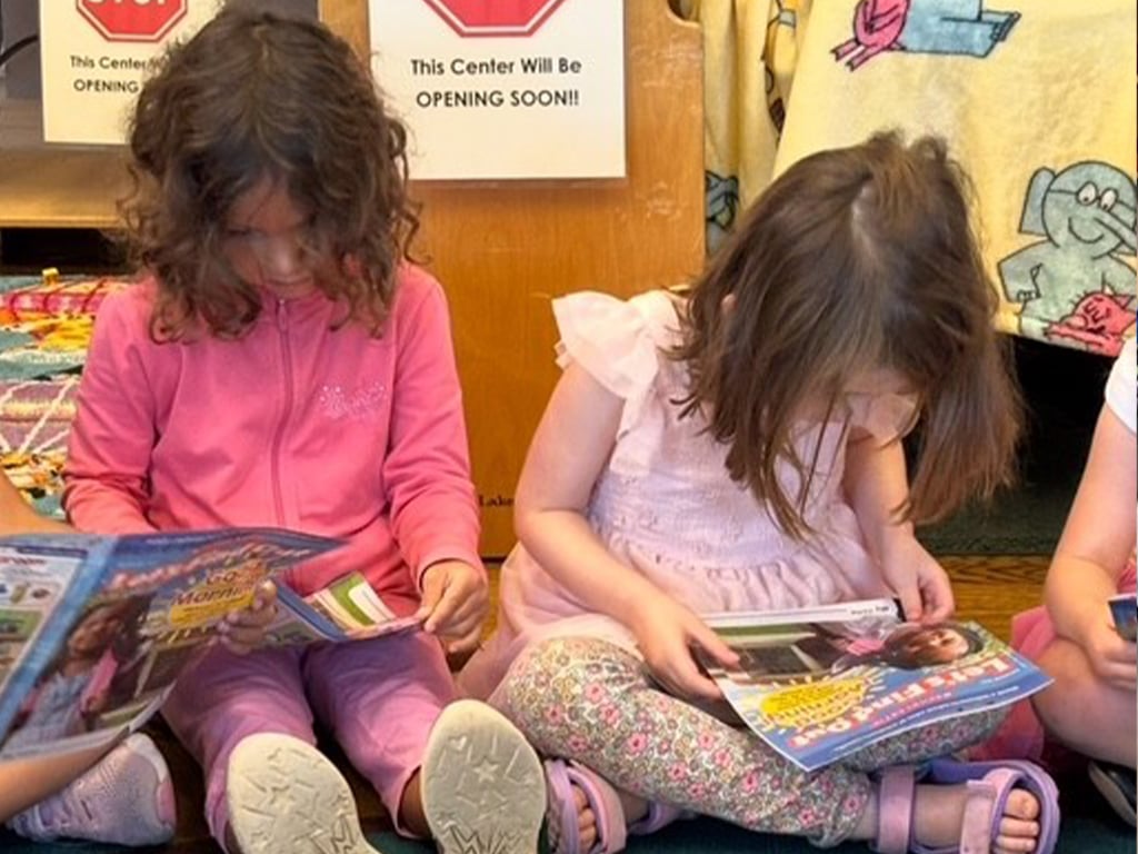 Two young girls sit on a floor and read magazines in a classroom setting