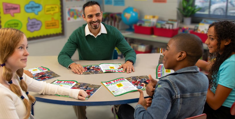 A man and three students sit at a round classroom table and read from open magazines together