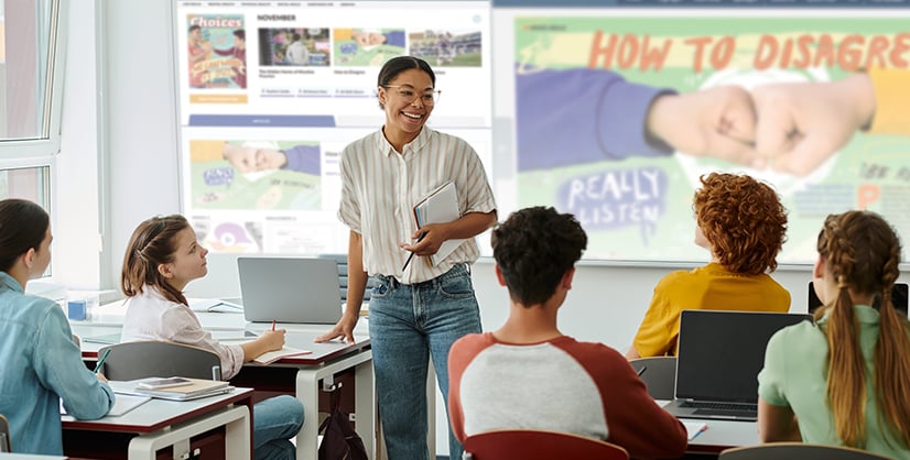 A teacher holds a laptop before students at desks as a screen behind her reads How To Disagree