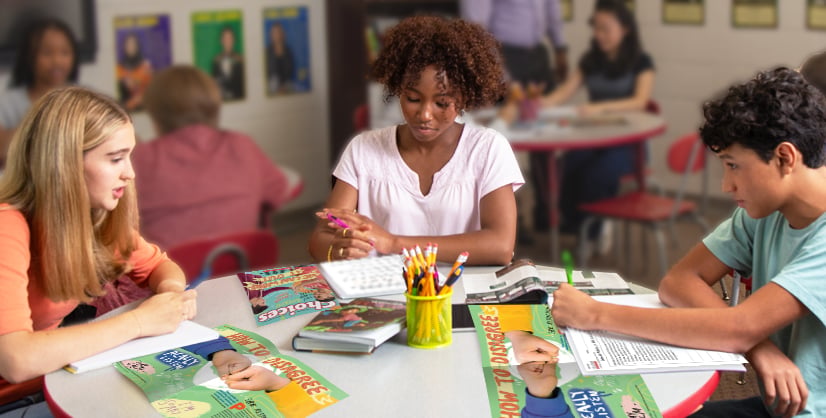 Three students sit around a table of booklets & a cup of pencils as the boy writes on a worksheet