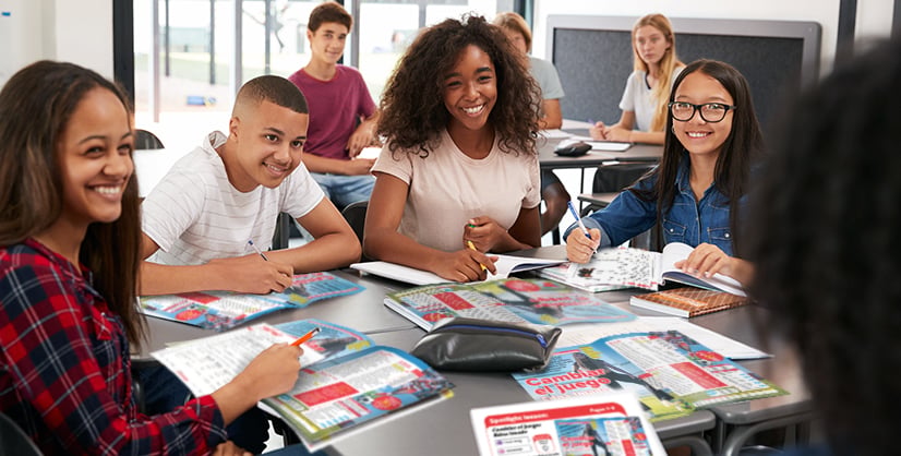 Four students sit together at a classroom table with open workbooks and notebooks