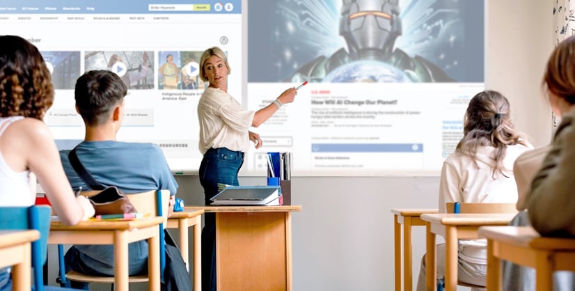A teacher points at a projected article reading How Will AI Change Our Planet? as 4 students watch