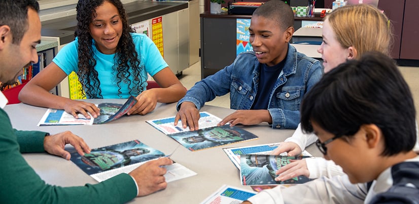 A teacher & 4 students sit around a classroom table, each turning the pages of a magazine