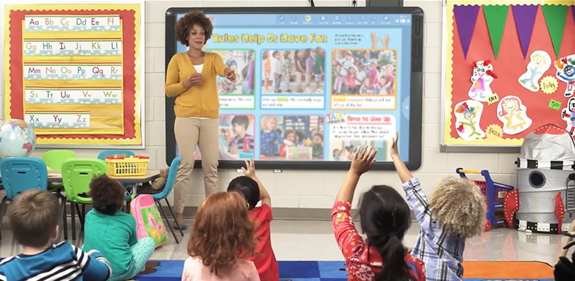 A woman leads a classroom lesson as young students raise their hands toward a smartboard