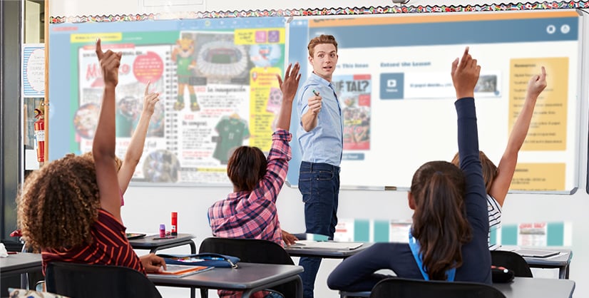 A teacher stands at a whiteboard as students raise their hands in a classroom