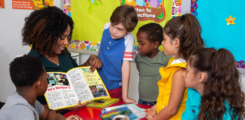 A woman reads an open magazine to a group of young children gathered around a classroom table