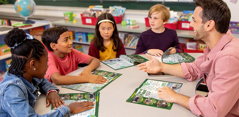 A man leads a small group of four students at a table with open magazines in a classroom