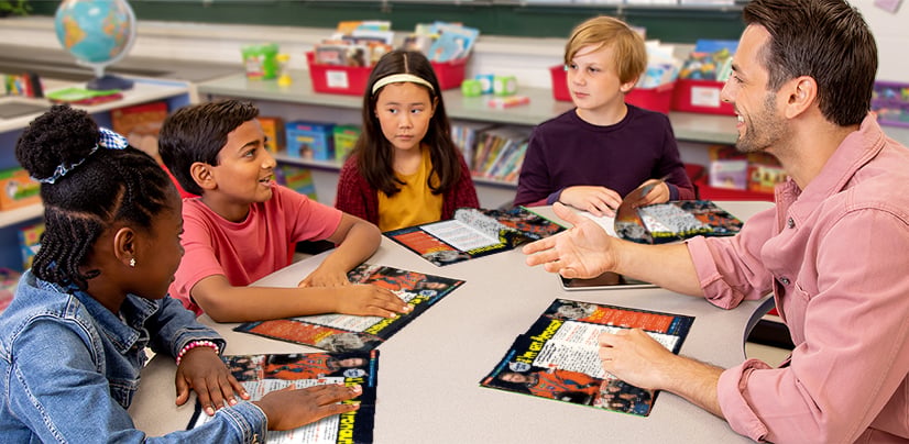 Four students and an adult sit around a table in a classroom and discuss printed materials