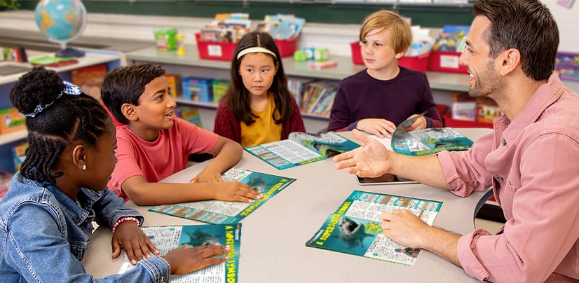 A man and four children sit around a table in a classroom and discuss open magazines