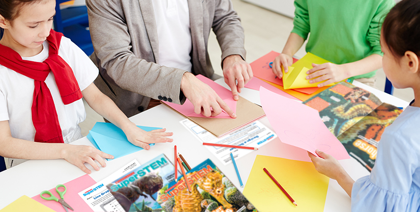 Three students and an adult work with colored paper and SuperSTEM magazines at a table