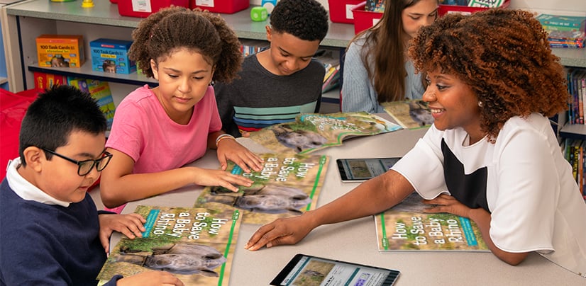 A woman and four students gather around a table with open books and tablets in a classroom