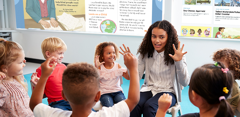 A woman leads a group of young children in an activity, all with hands raised, in a classroom