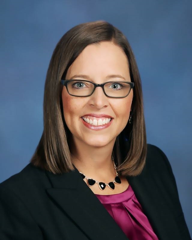 A professional headshot of a woman who wears glasses and a dark blazer