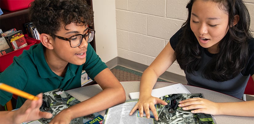 Two students sit at a table and discuss an open book together